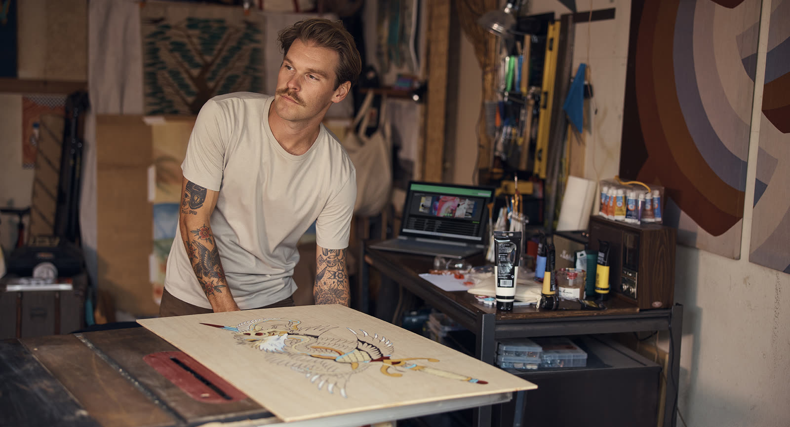 A tattooed man at a workbench studying a layout