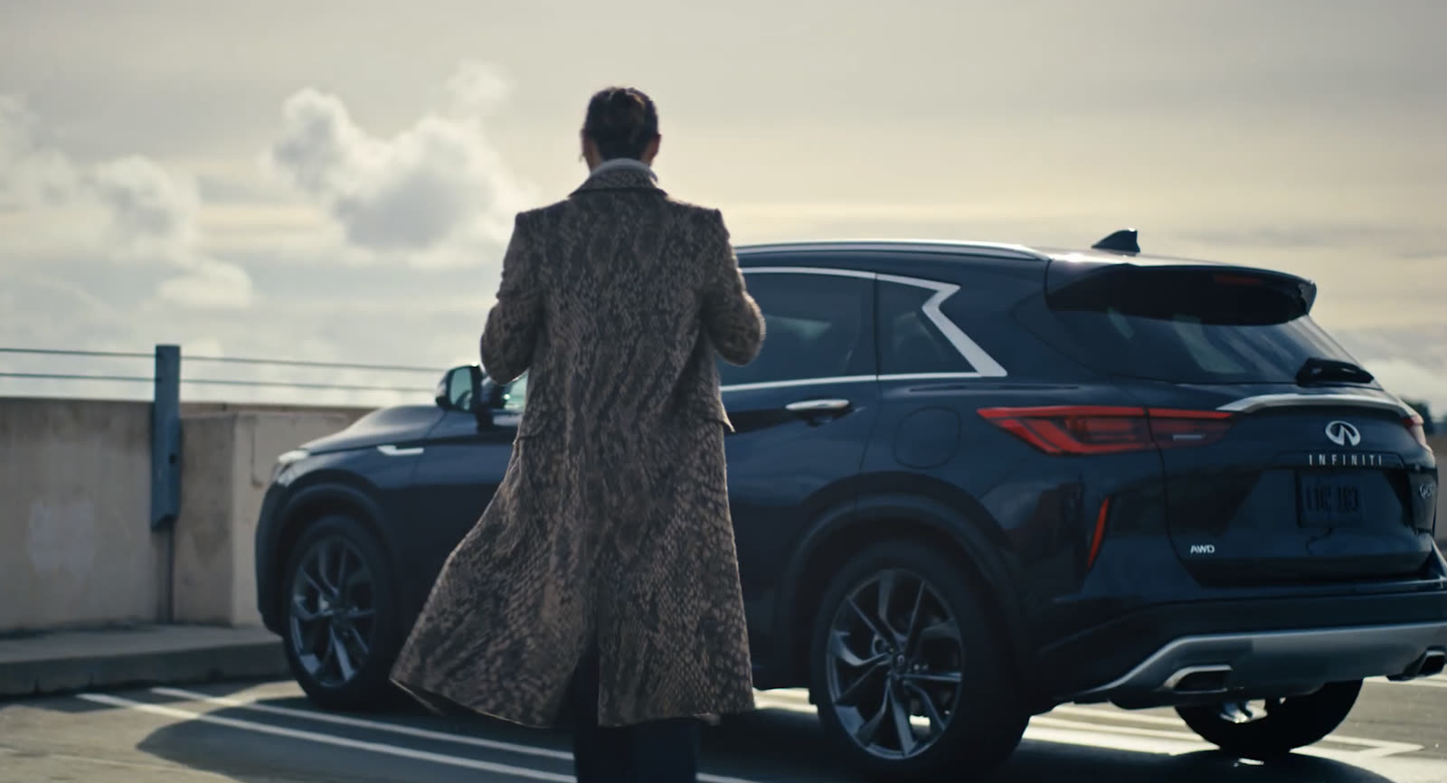 A figure in a long coat walking toward a dark Infiniti on a rooftop lot
